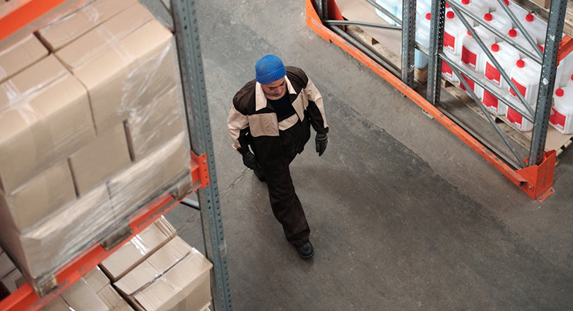 Worker walking through warehouse aisle surrounded by pallets and shelves with products.