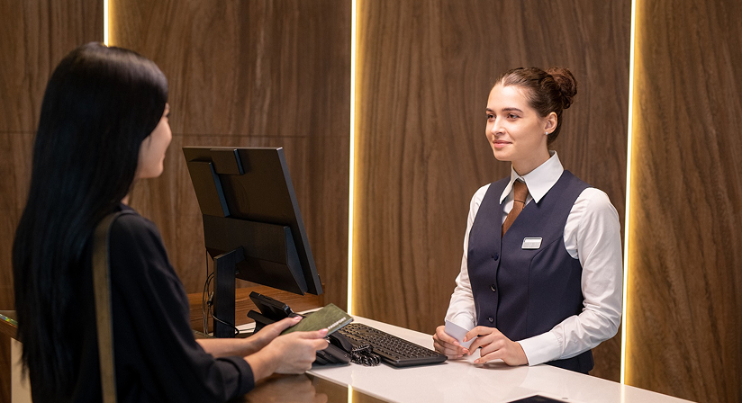 Hotel receptionist assisting guest at check-in counter, with modern decor in the background.