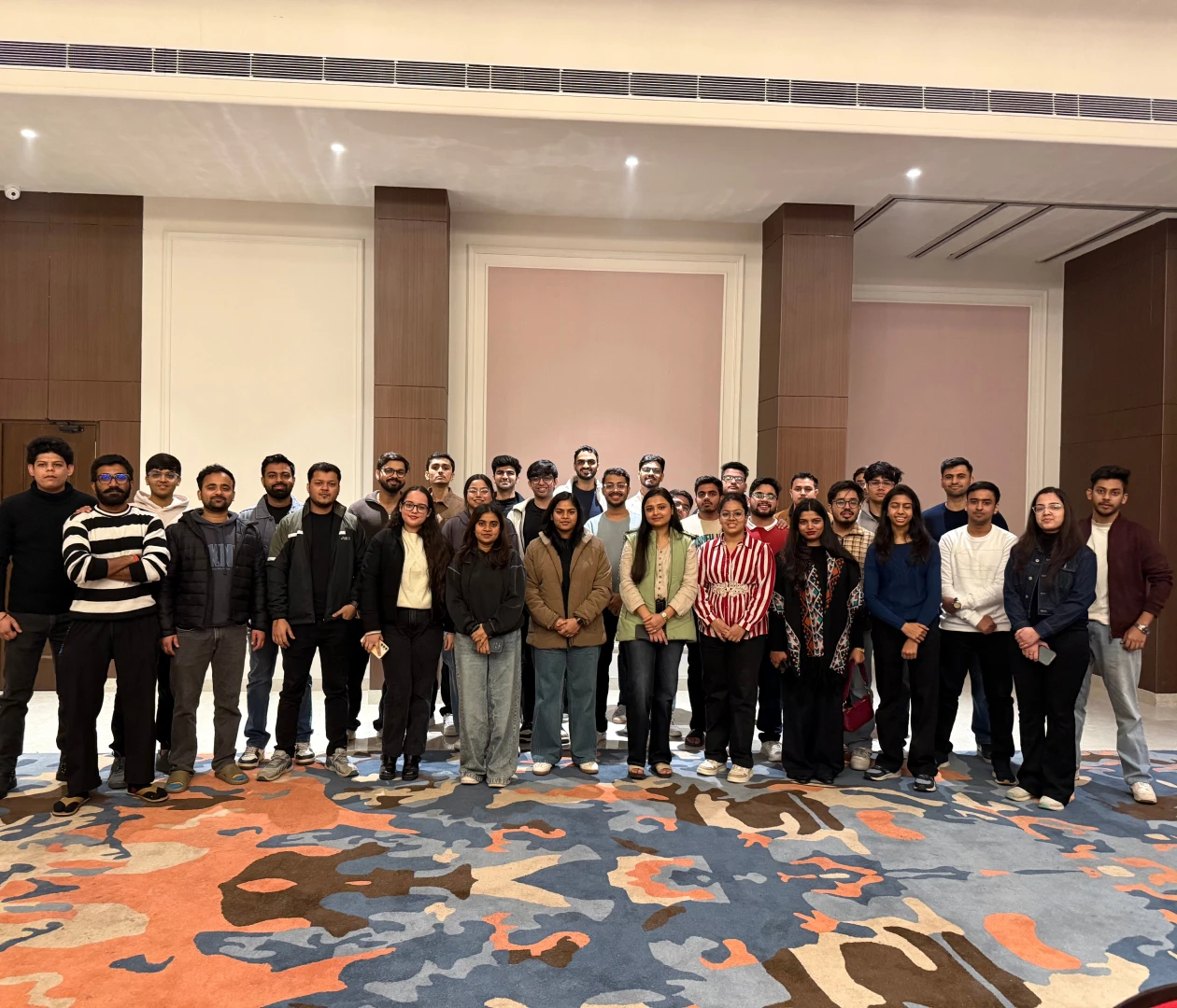 Group of people standing in a room during a gathering, colorful carpet visible below.