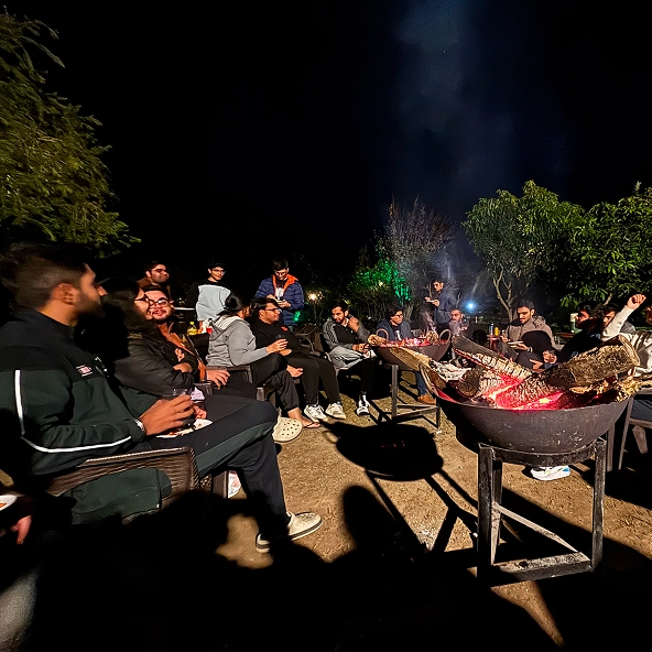Group of friends enjoying a night bonfire outdoors, surrounded by trees, sharing conversation and warmth.