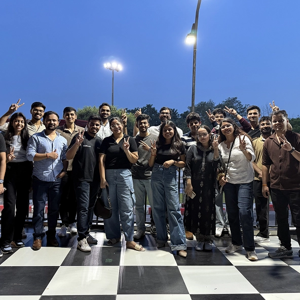 A group of people smiling and posing on a checkered floor at night, showing peace signs under bright streetlights.