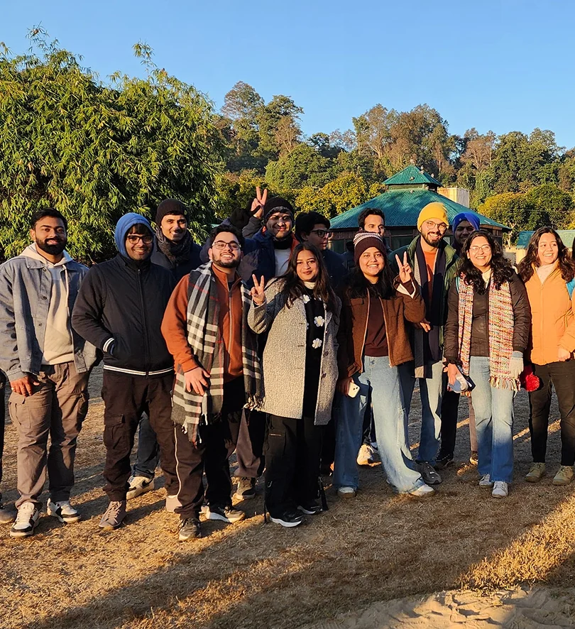Group of people outdoors in winter clothing, enjoying a sunny day with trees and a building in the background.