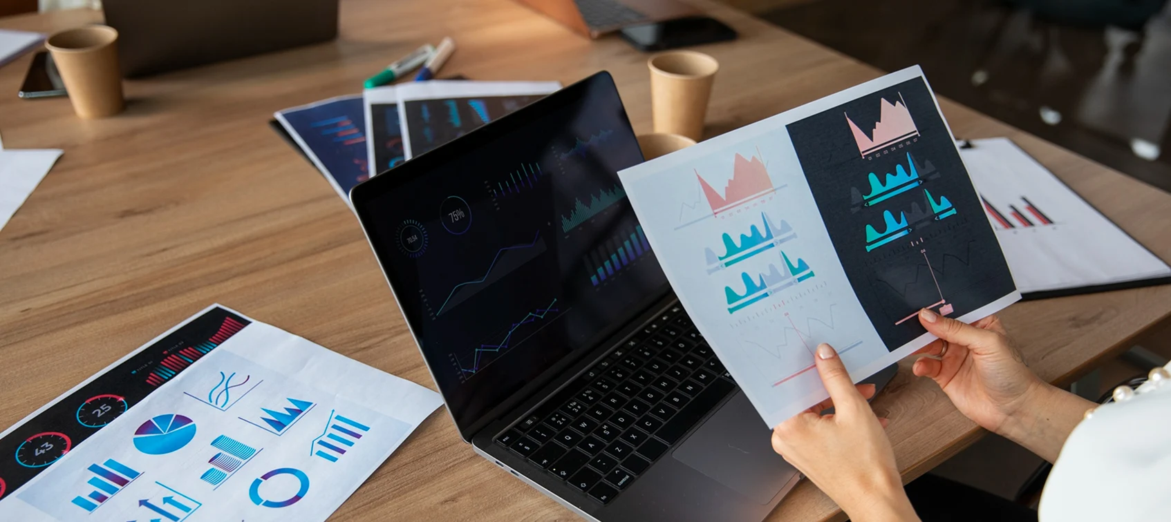 Person analyzing graphs and charts on a laptop and printed papers at a wooden desk.